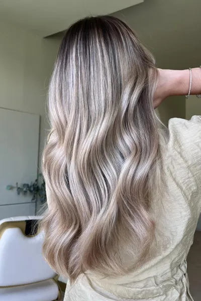 Balayage blond beige froid sur cheveux longs ondulés, racines fondues et longueurs lumineuses avec reflets naturels